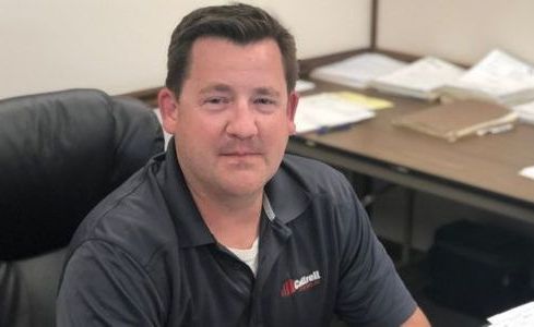 Man seated at an office desk in a dark polo shirt, looking at the camera with papers and folders behind him.