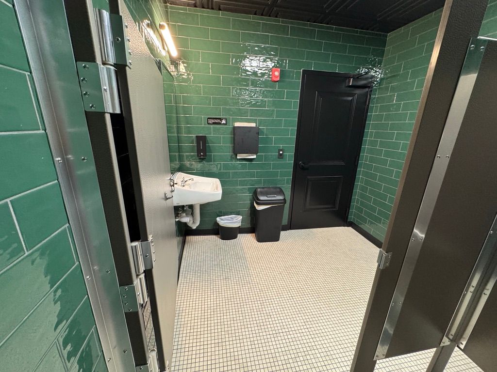 Green-tiled public restroom with sink, toilet, trash can, and a black door opening into a stall