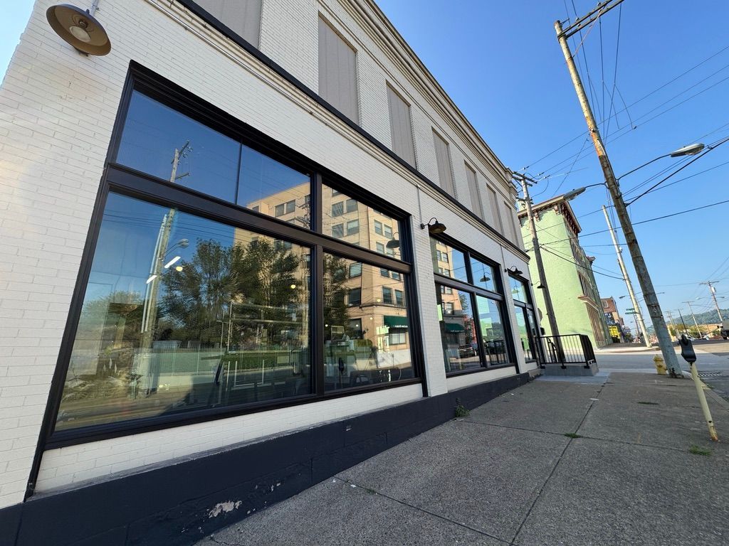 Street-level view of a modern storefront with large black-framed windows on a sunny sidewalk.