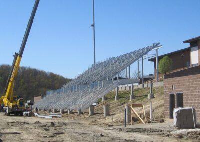 Crane lifting a large metal bleacher frame at a construction site beside a brick building.