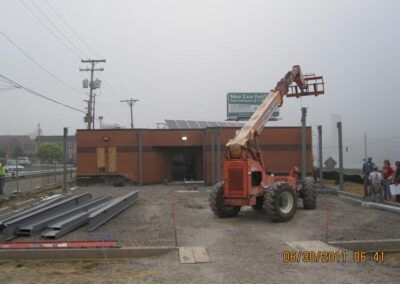 Orange construction lift in front of a brick building at a muddy worksite, with stacked metal beams nearby