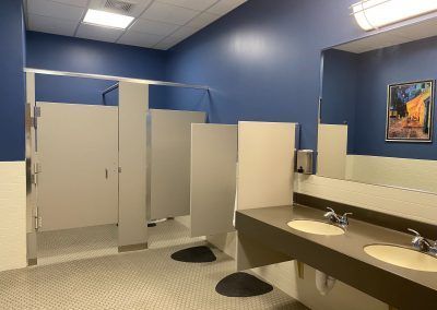 Public restroom with blue walls, white stalls, and beige sinks under a framed picture.