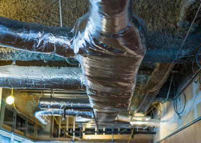 Insulated HVAC ductwork and exposed pipes in an industrial ceiling with fluorescent lights