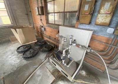 Industrial utility room with exposed pipes, gauges, and coiled hoses against a brick wall.