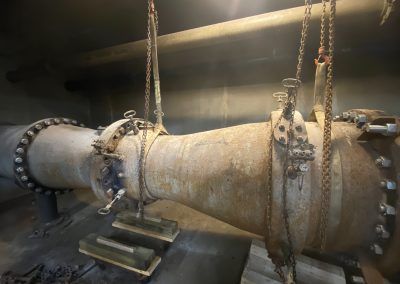 Large rusted cylindrical industrial pipe suspended by chains in a dim metal chamber