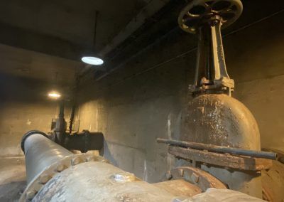 Industrial pumps and large pipes in a dim concrete room with water spraying from a valve.