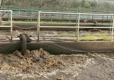 Mud and waste flowing from a pipe into a muddy ditch beside a fenced industrial area