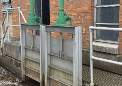 Metal flood barriers installed beside a brick building with green pipes and handrails