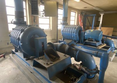 Two large blue industrial pumps in a workshop, connected by pipes and mounted on metal bases