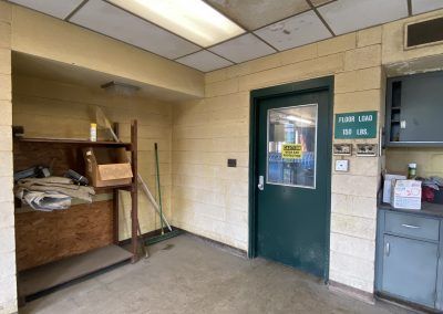Small utility room with shelves, green door, and gray vending machine against beige cinderblock walls.