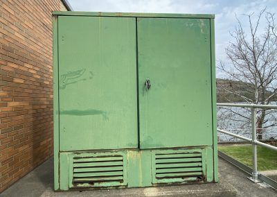 Green metal utility cabinet with vented bottom doors beside a brick wall outdoors