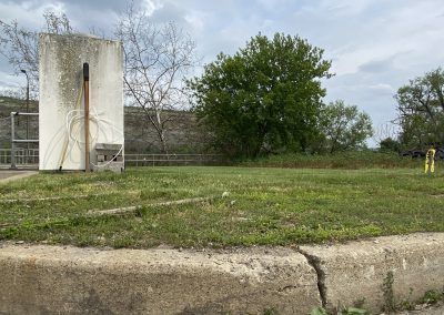 Grassy field beside a concrete wall with a hose and a distant yellow fire hydrant under cloudy skies