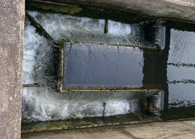 Top-down view of a narrow concrete water channel with churning white water around a dark rectangular pool.