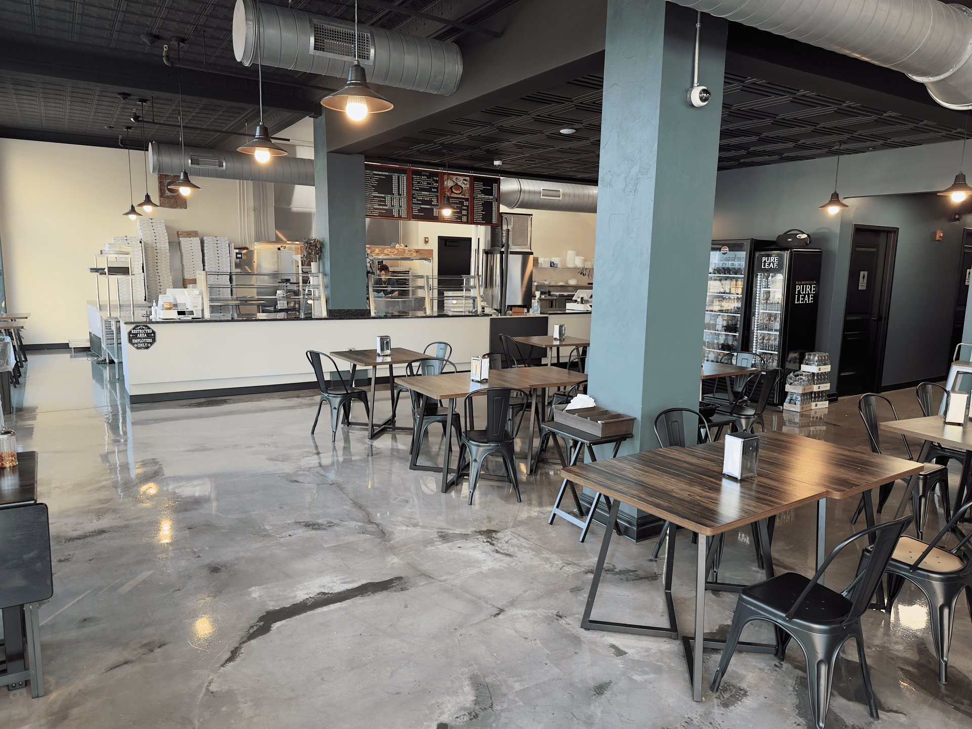 Empty modern café interior with metal tables, black chairs, and a white counter under exposed ductwork