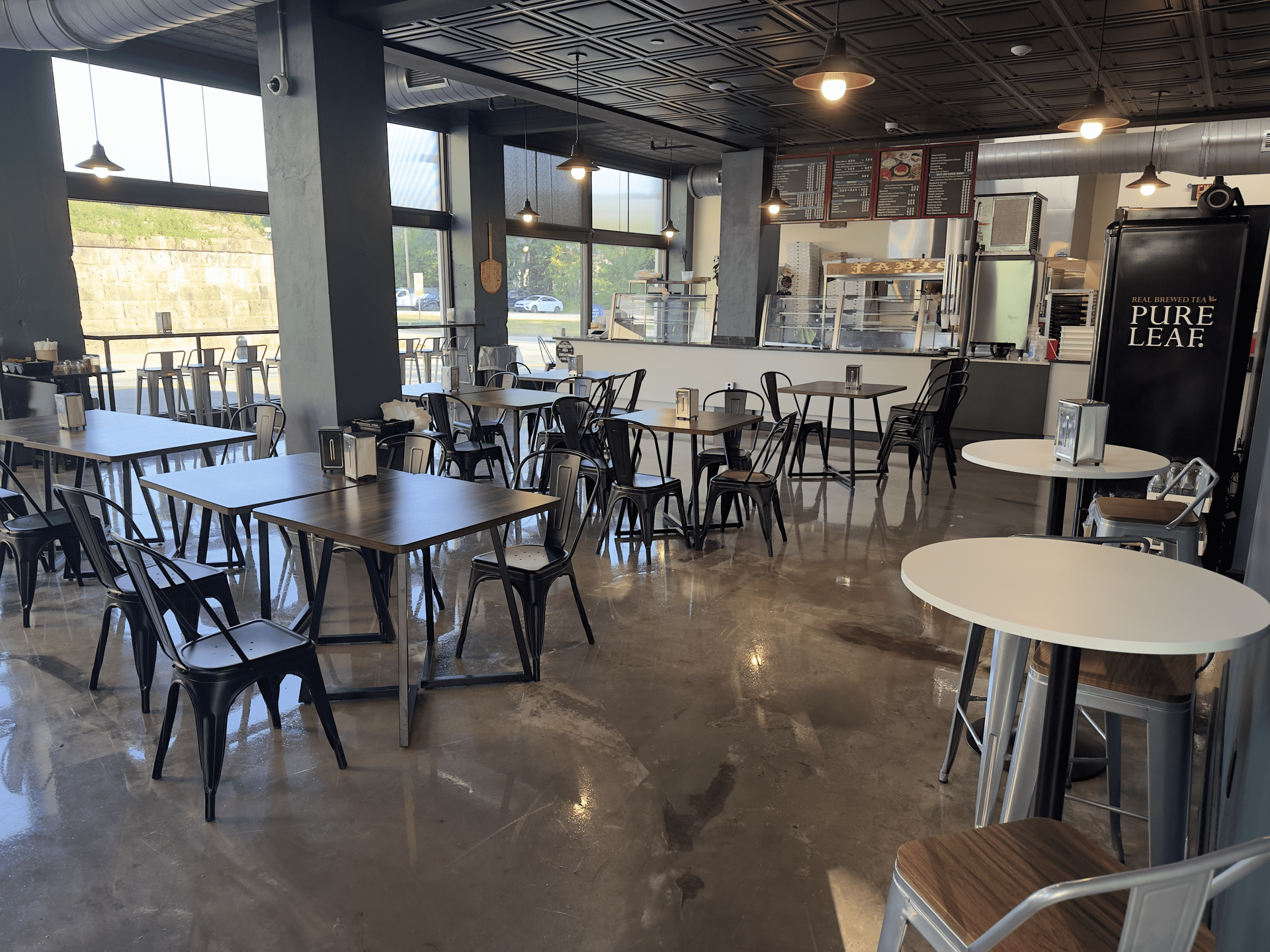 Empty café with black tables and chairs, large windows, and a polished concrete floor.