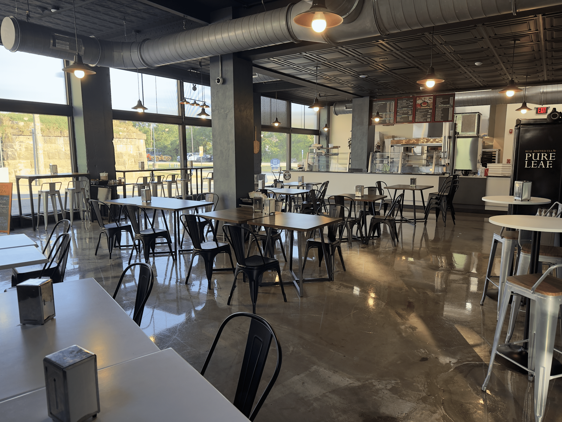 Empty modern café seating area with metal tables and chairs, polished concrete floor, and large windows.