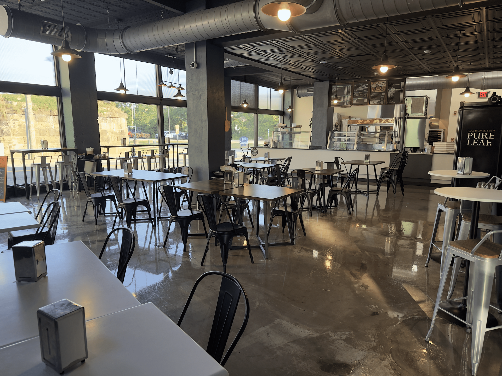 Modern café interior with metal tables, stools, and chairs, lit by large windows and hanging lights.