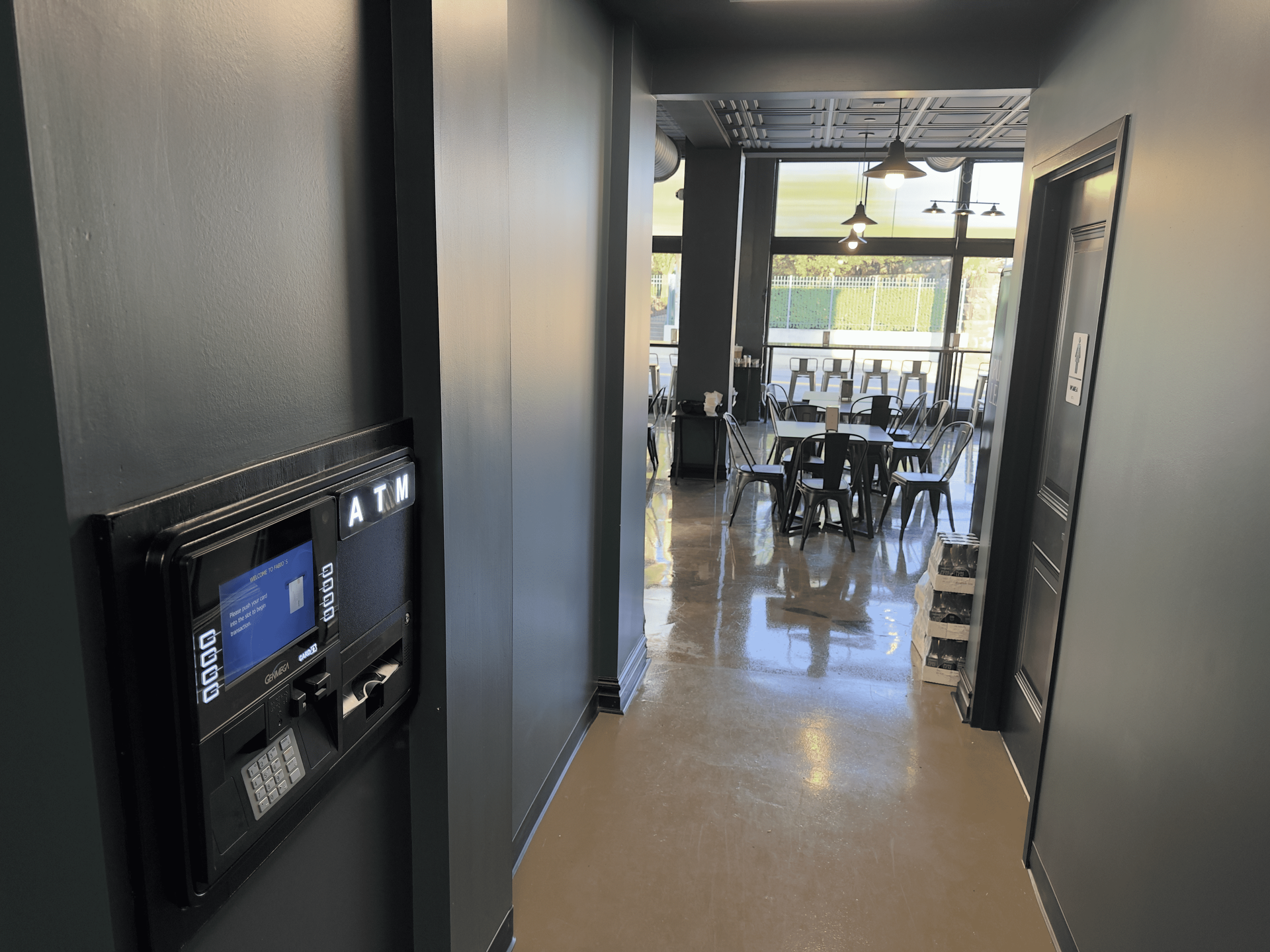 Hallway with a wall-mounted control panel leading to a bright dining area with tables and chairs.