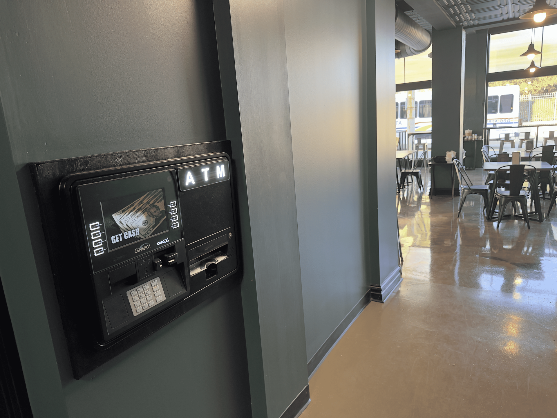 Wall-mounted touchscreen kiosk in a gray hallway, with a cafeteria seating area visible in the background