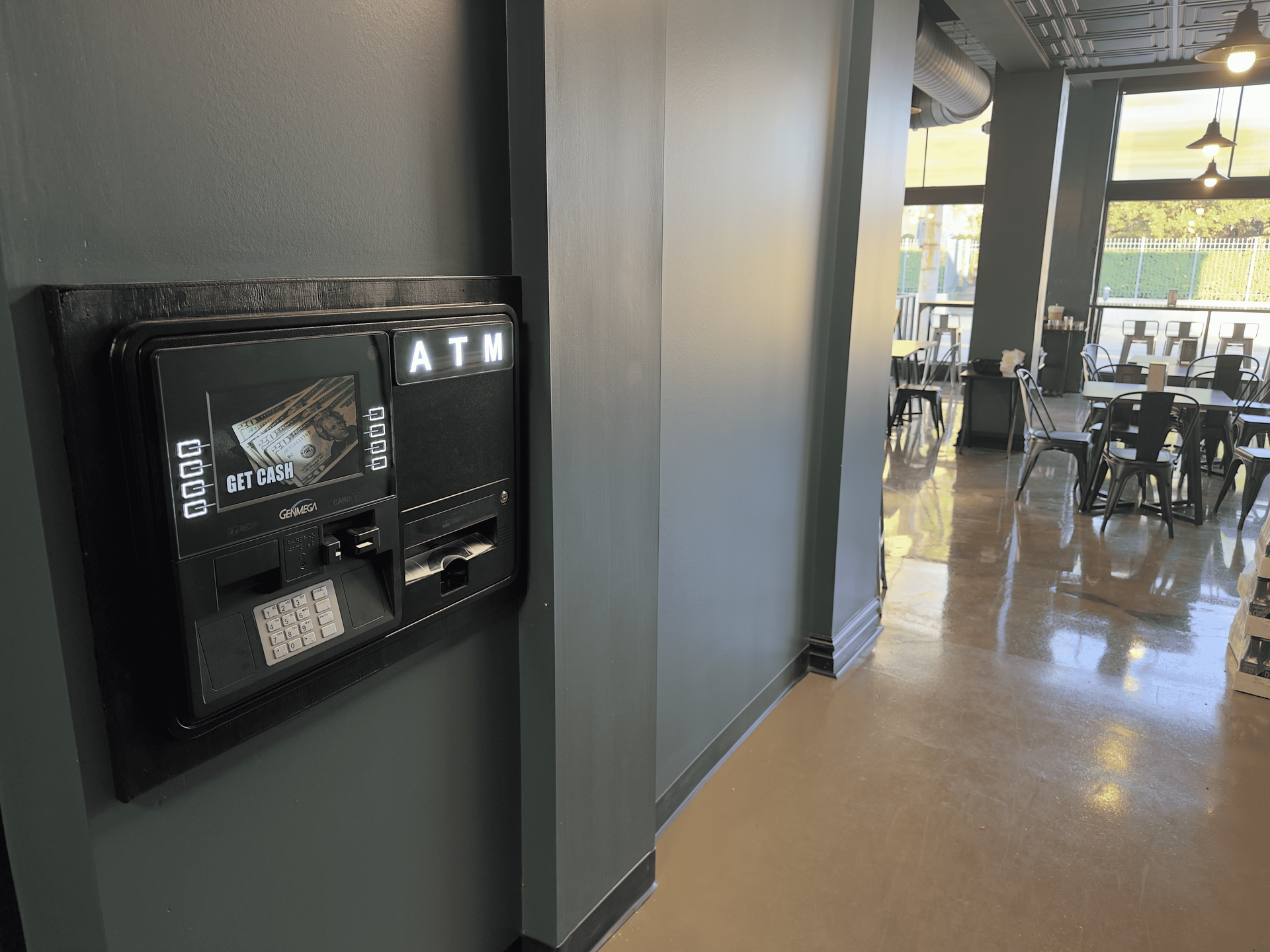 Wall-mounted ATM on a dark gray wall in a bright lobby or café area
