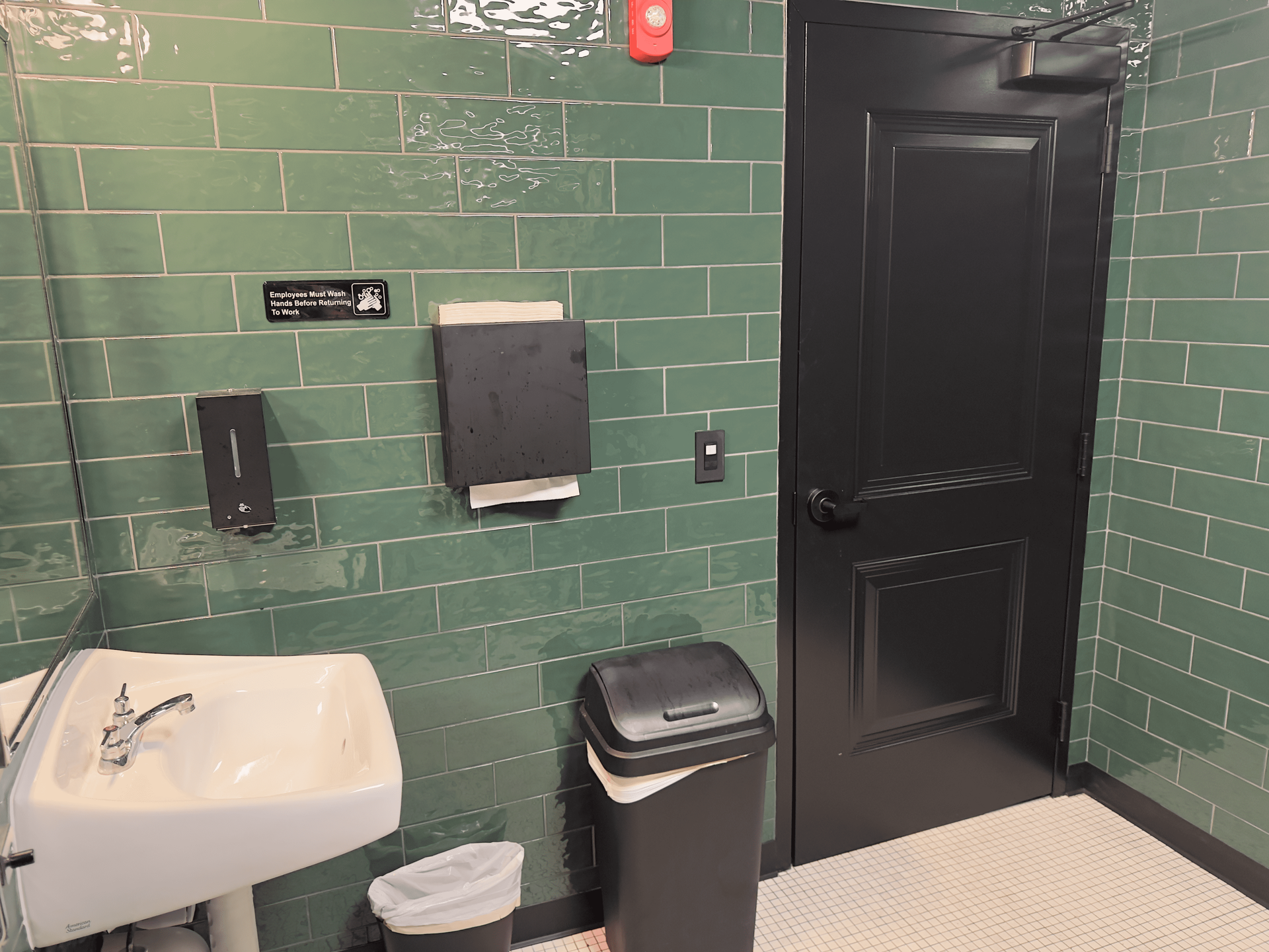Green-tiled restroom with sink, paper towels, trash can, and black door