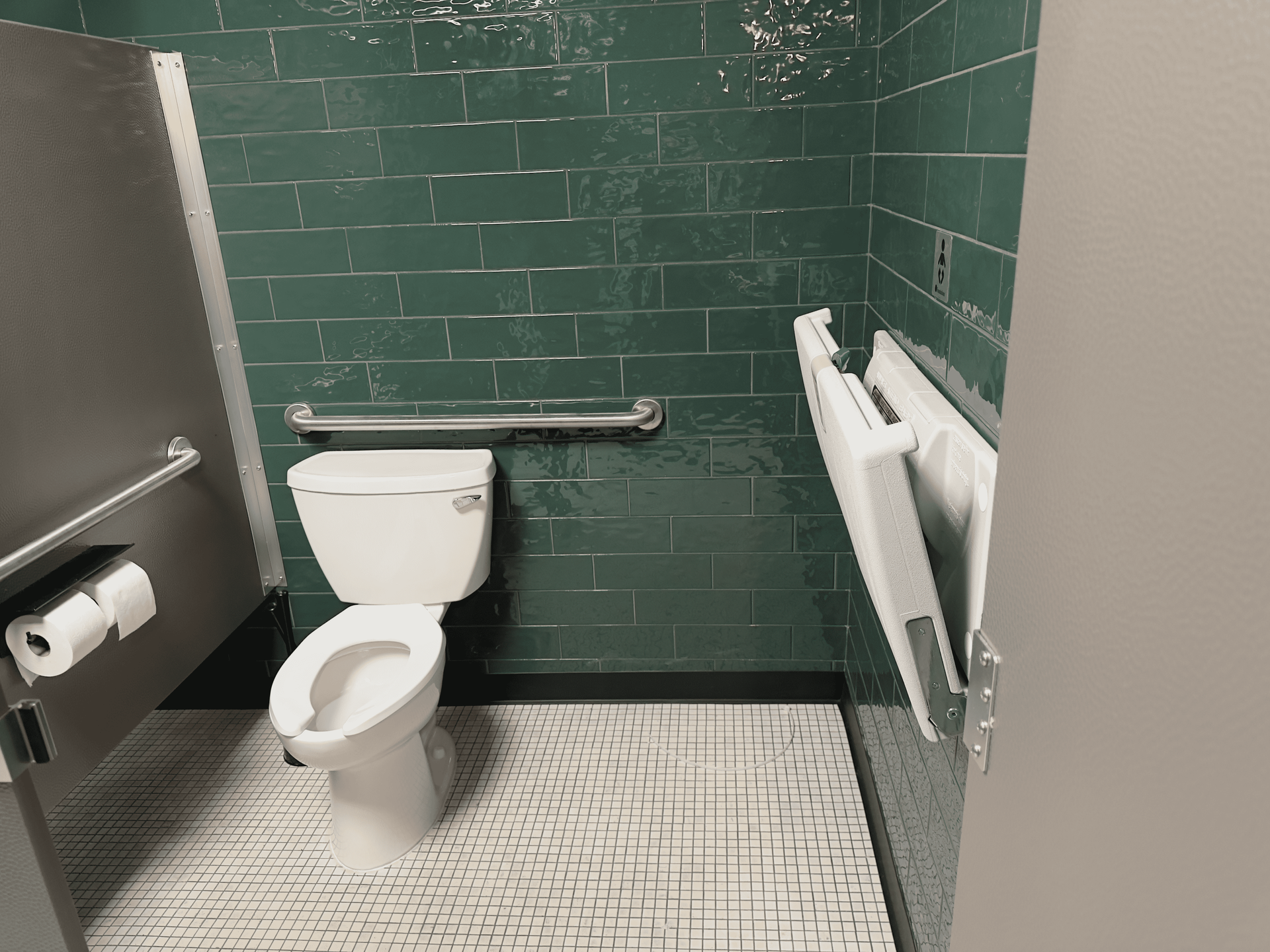 Accessible restroom with toilet, grab bars, and wall-mounted changing table in green-tiled room