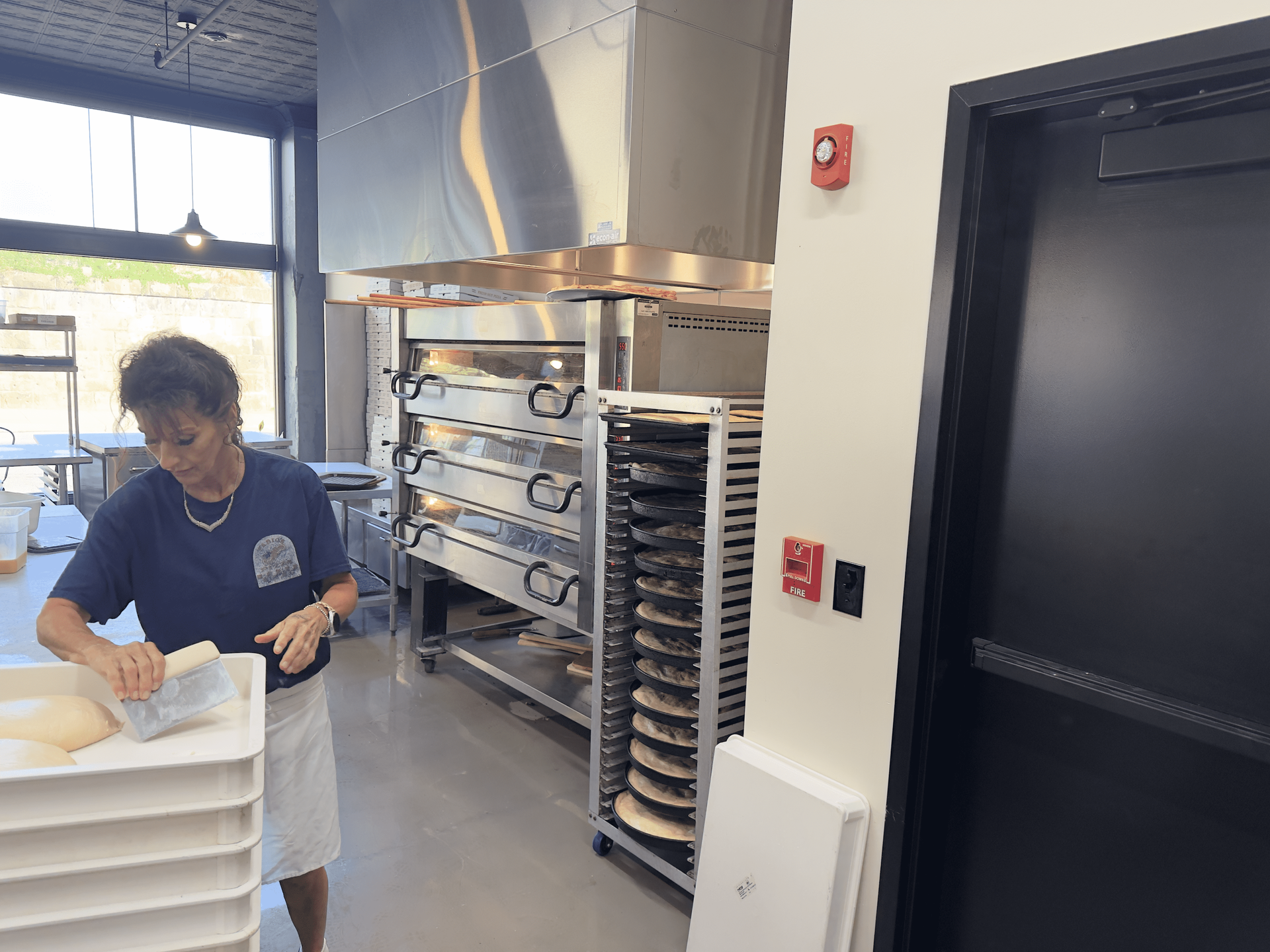 Worker stacking white trays in a commercial kitchen with stainless steel equipment and open ovens