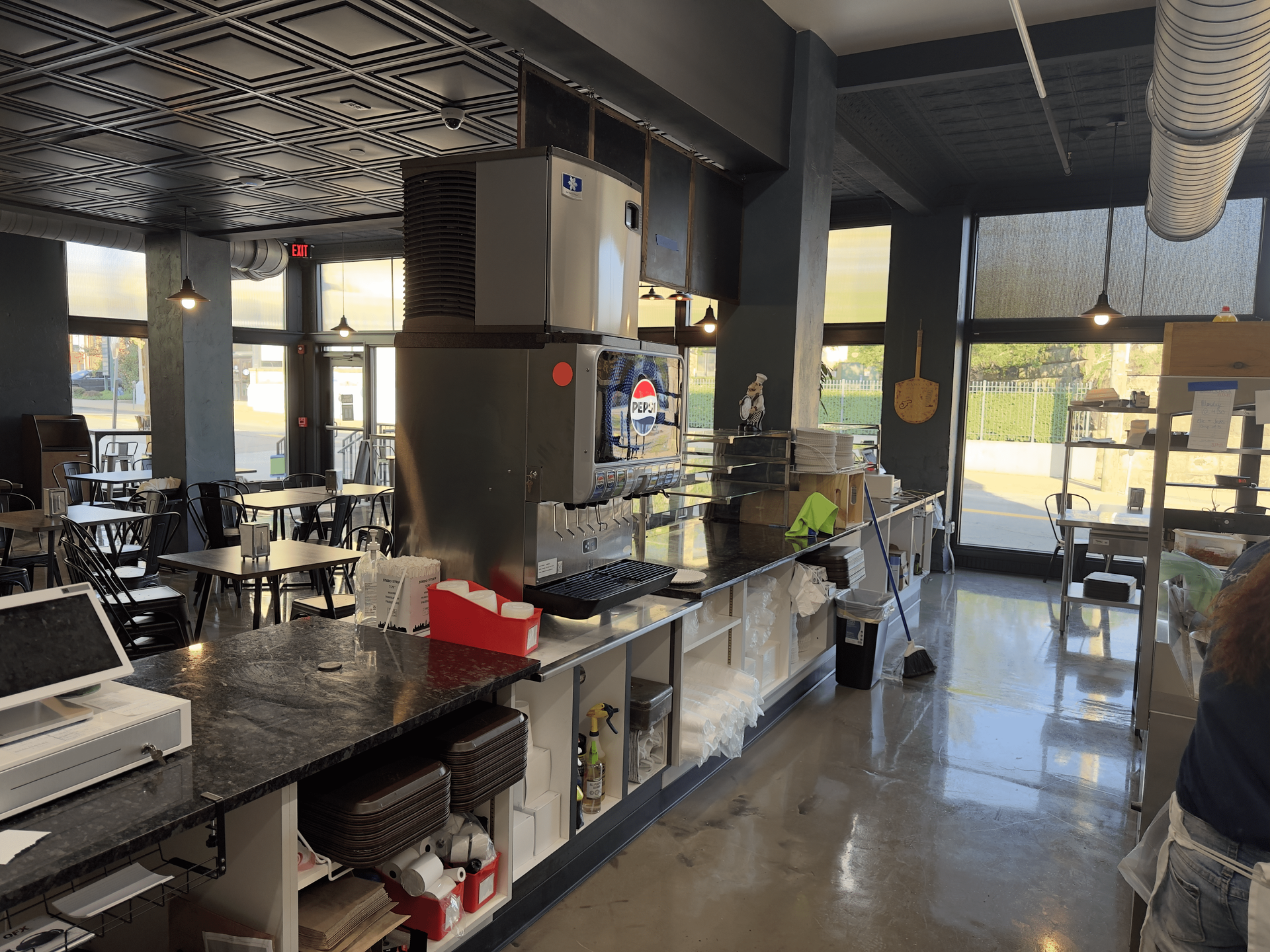 Empty café counter with drink station, tables, and sunlight streaming through front windows.