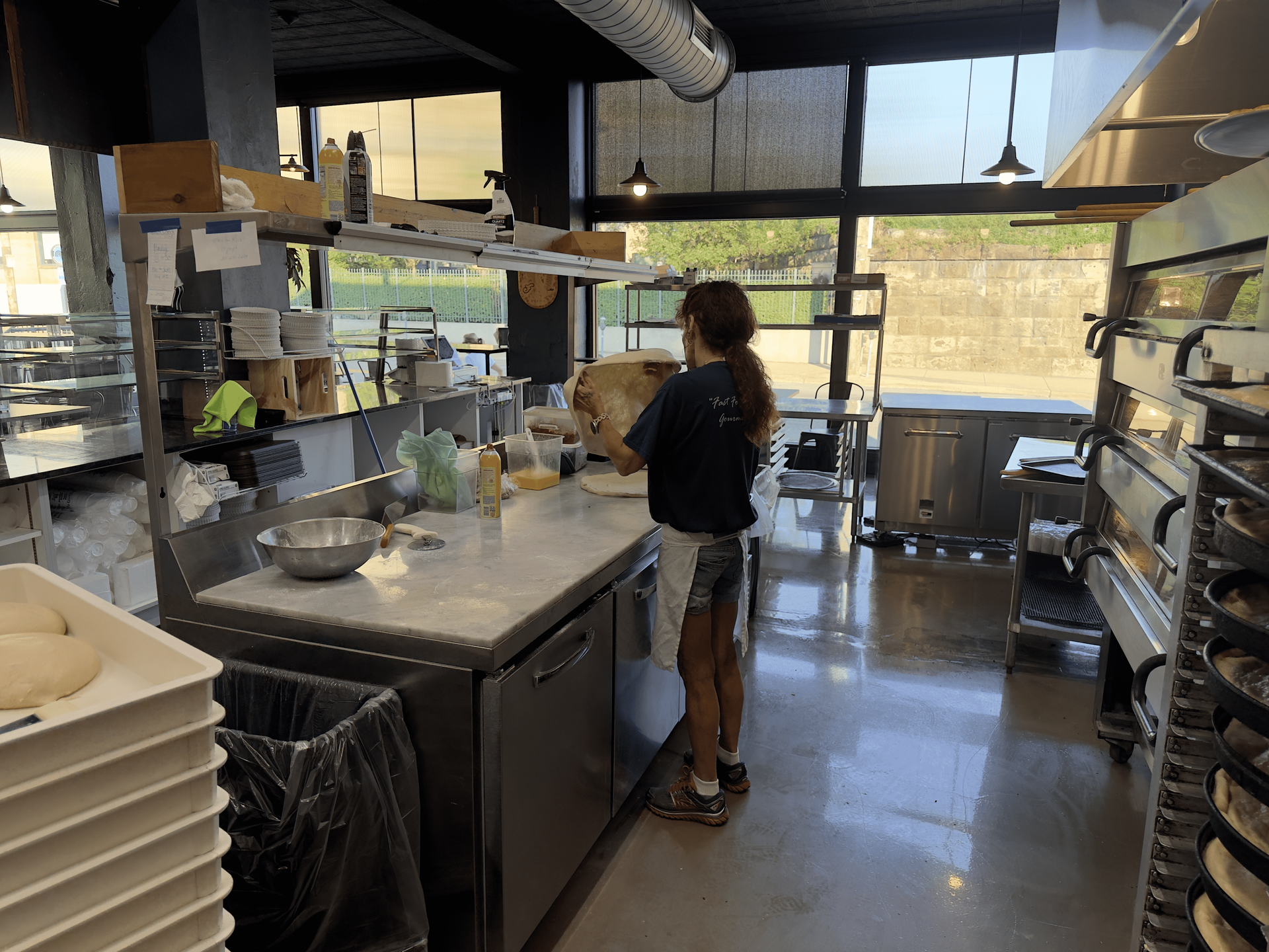 Person working in a bright commercial kitchen beside shelves, sinks, and stacked trays.