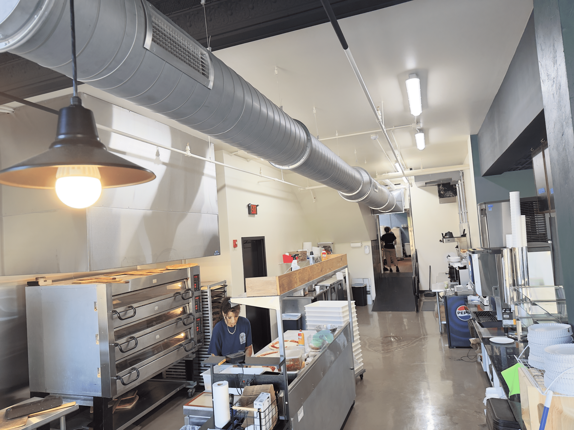 Industrial kitchen with stainless steel ovens, ventilation ducts, and prep counters along a narrow aisle