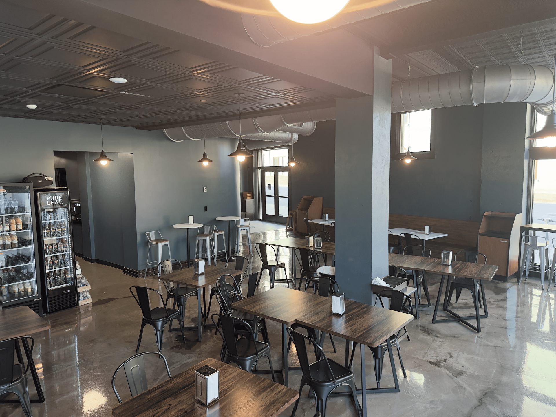 Empty modern café with tables and chairs, dark walls, reflective floor, and display fridge near the counter.