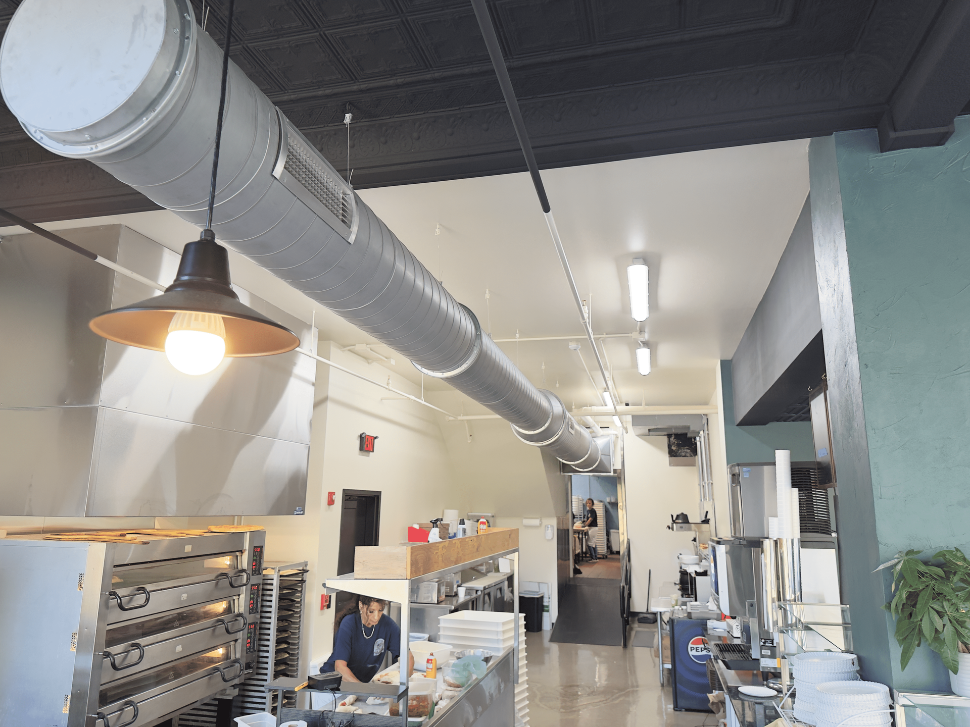 Industrial kitchen or bakery interior with stainless equipment, shelves, and large ventilation ducting under bright lights