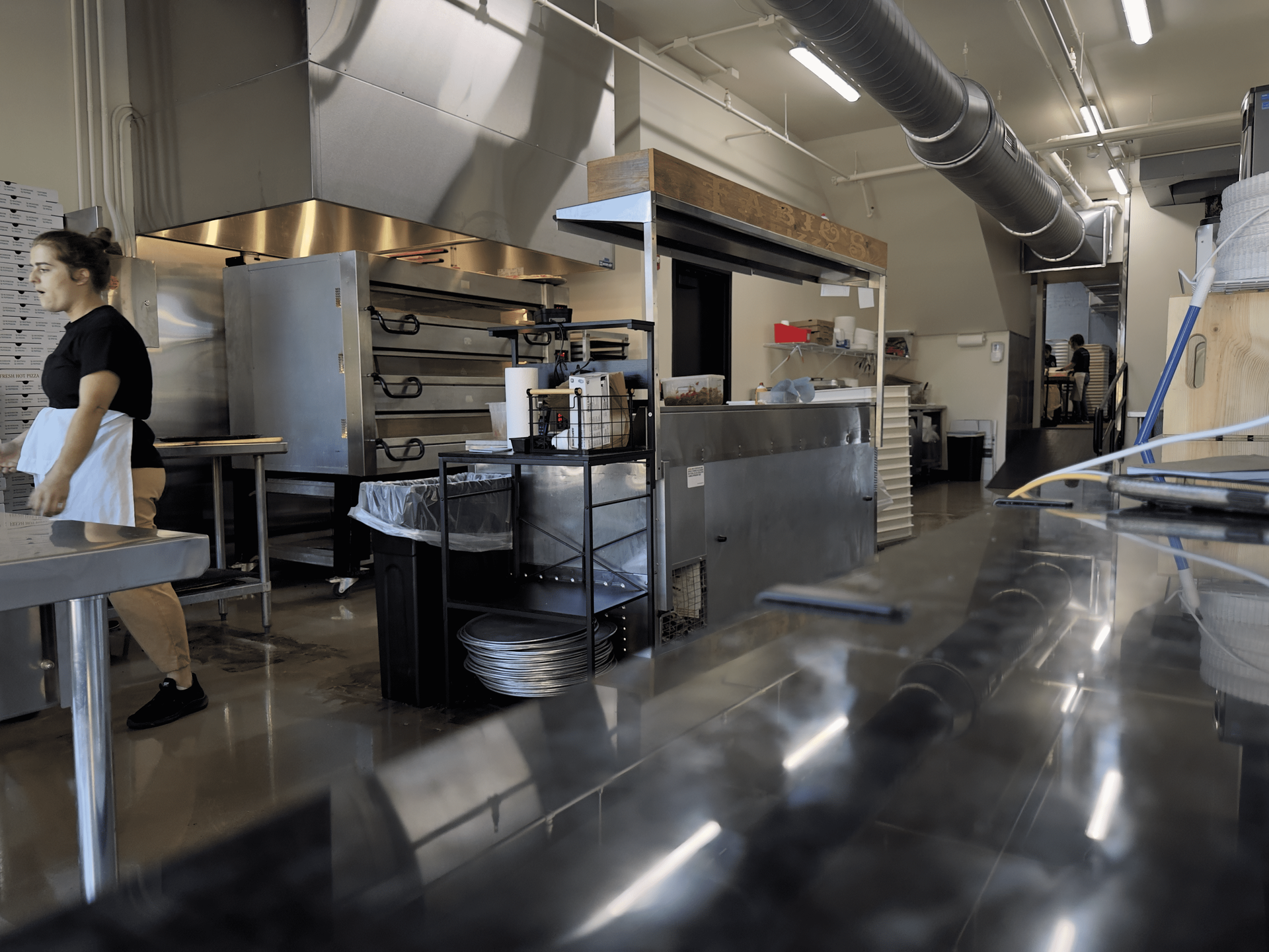 Commercial kitchen with stainless steel counters, ovens, vents, and a worker in an apron on the left