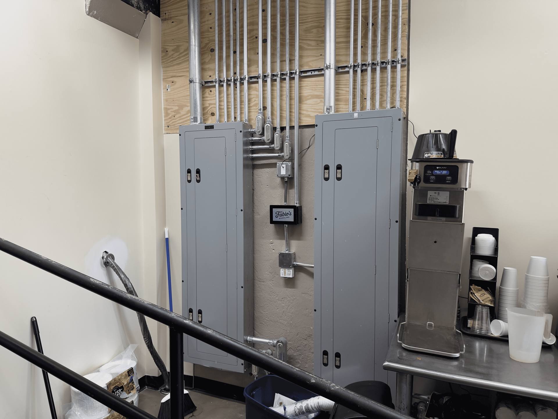 Electrical room with gray cabinets, conduit pipes, and a small sink beside a black railing.