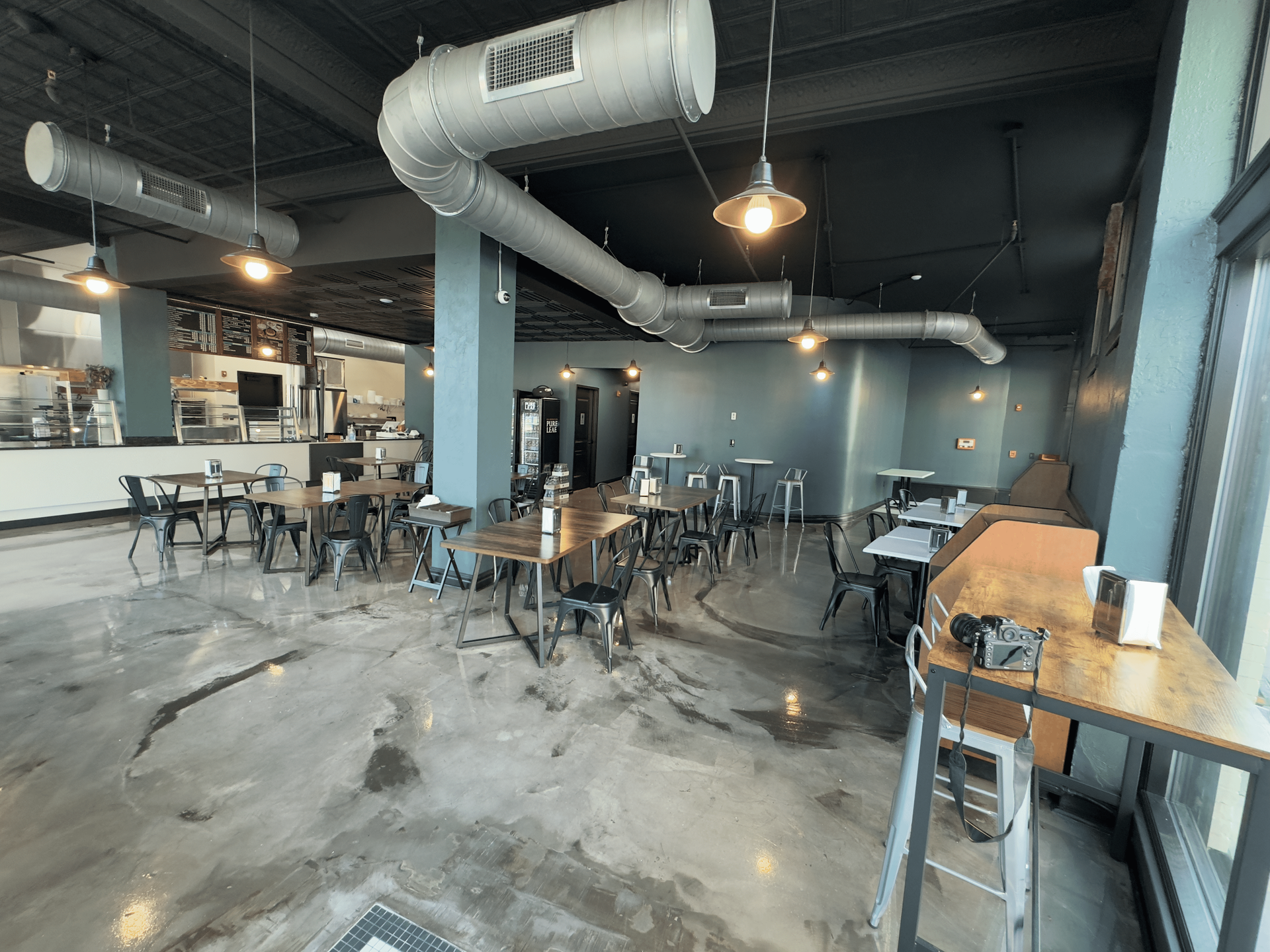 Empty industrial-style cafe with metal tables, hanging lights, and exposed ductwork in a gray interior.