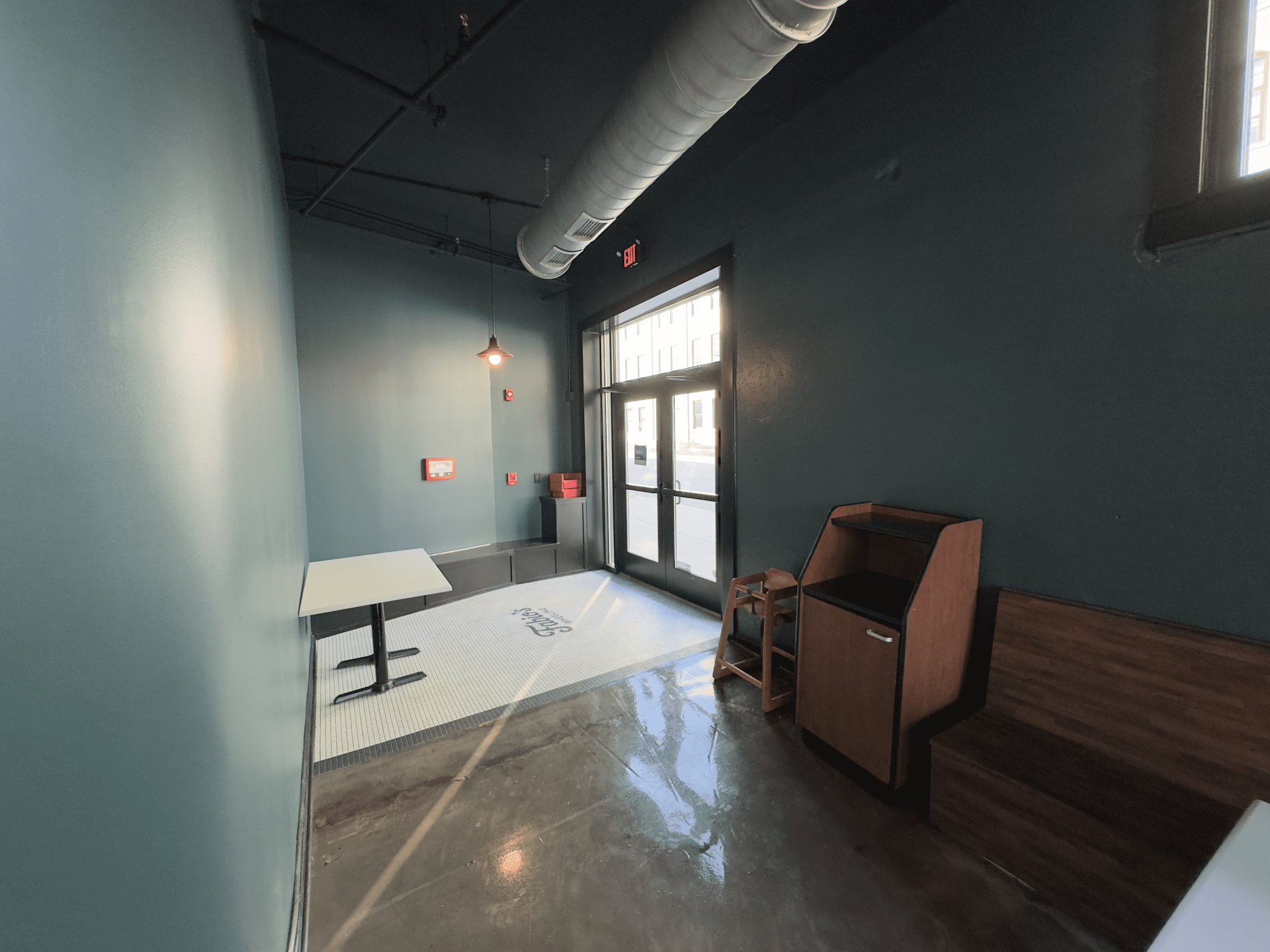 Modern lobby with teal walls, glass door, table, chair, and brown bench seating under exposed ductwork.
