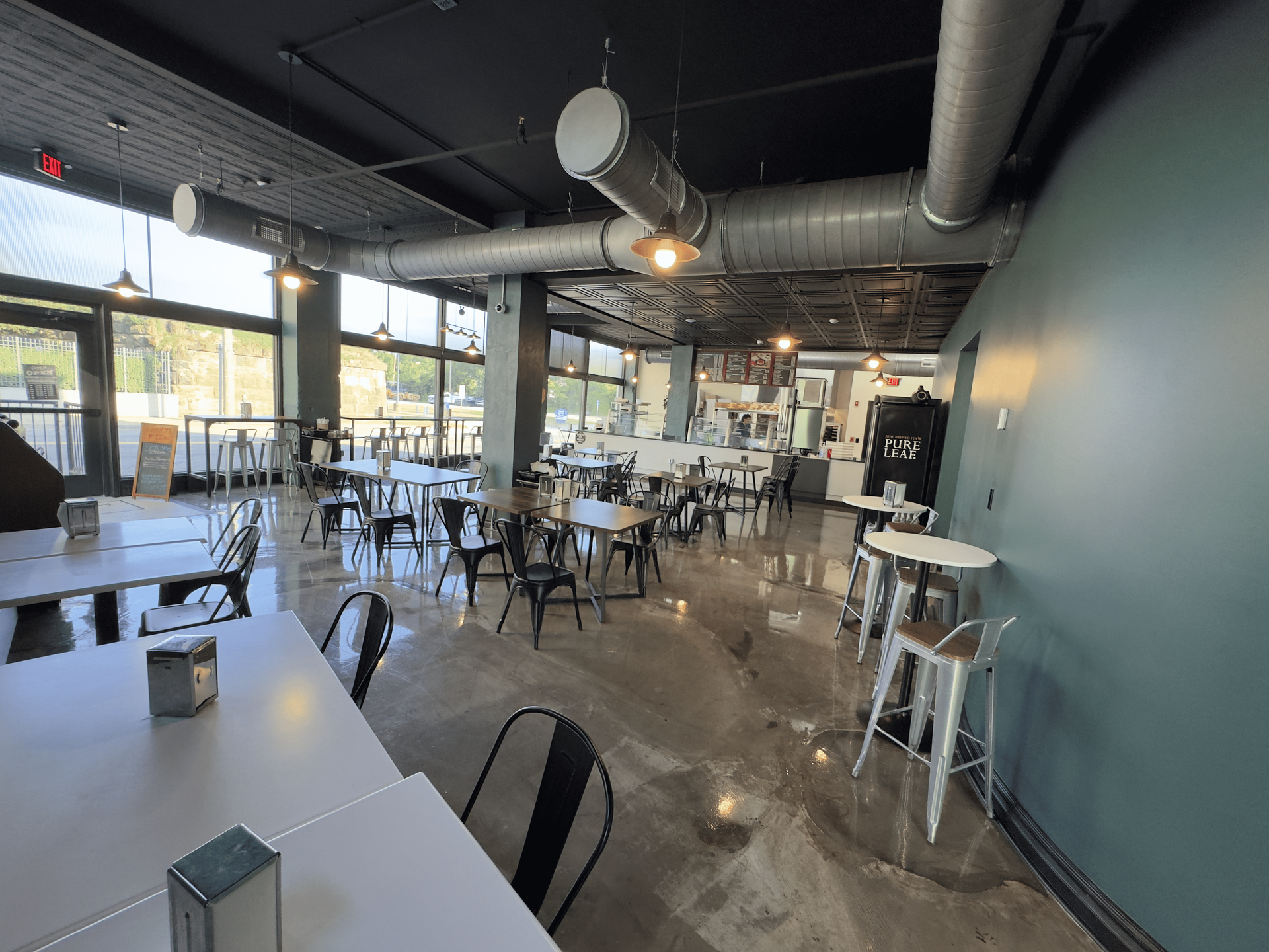 Empty modern cafe dining area with tables, chairs, and large windows under exposed ductwork.