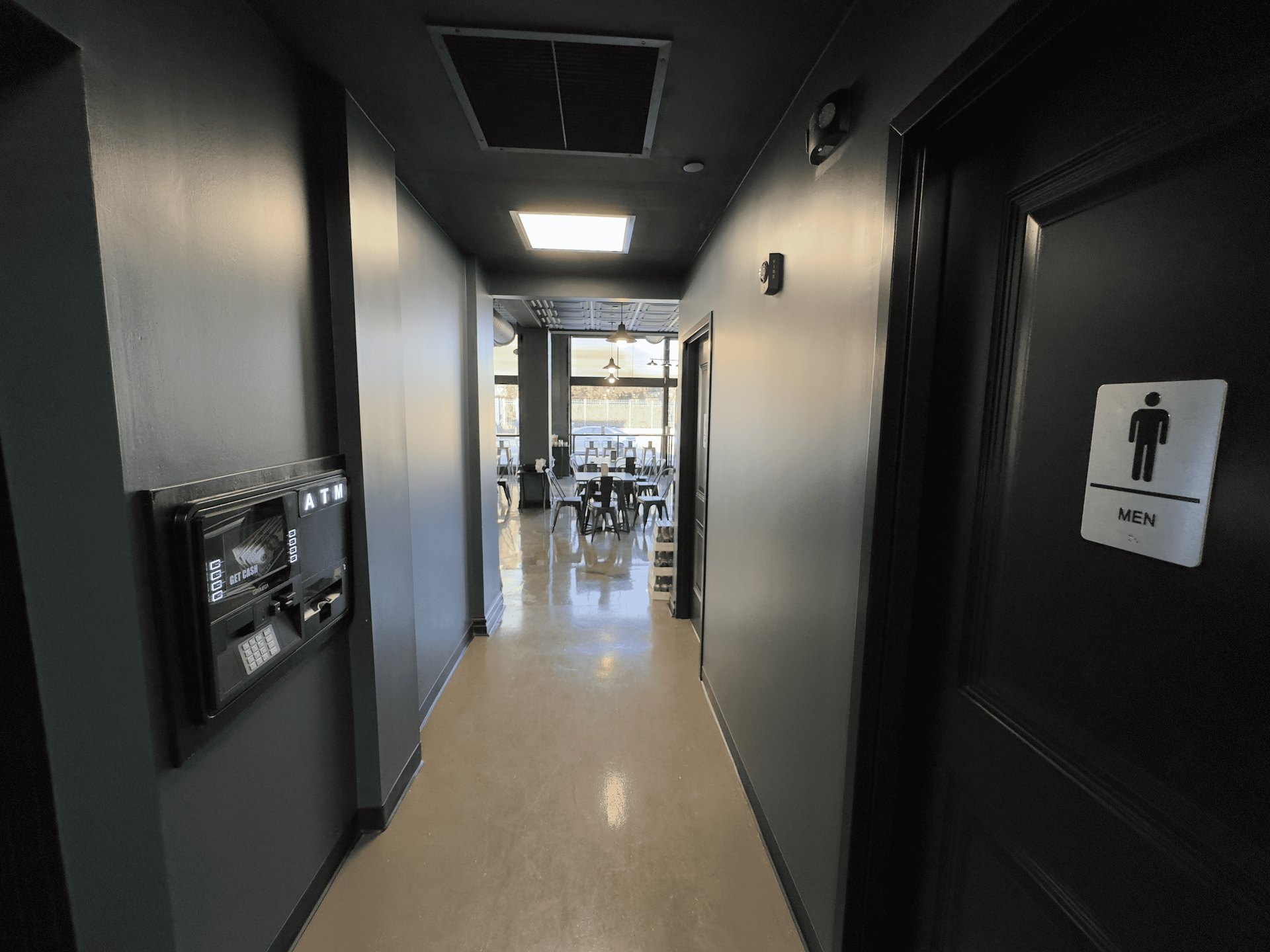 Narrow hallway with dark walls and beige floor leading to a brighter room with tables and chairs.