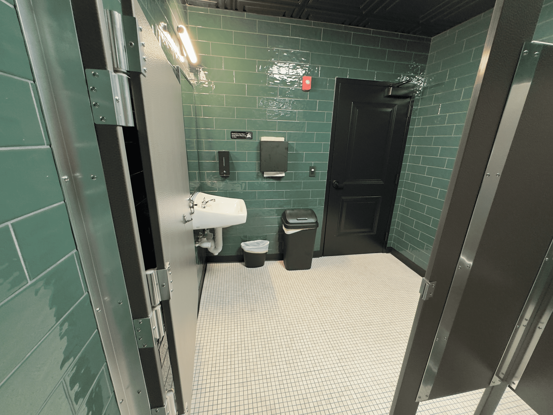 Green-tiled restroom stall with white sink, toilet, trash can, and black stall door.
