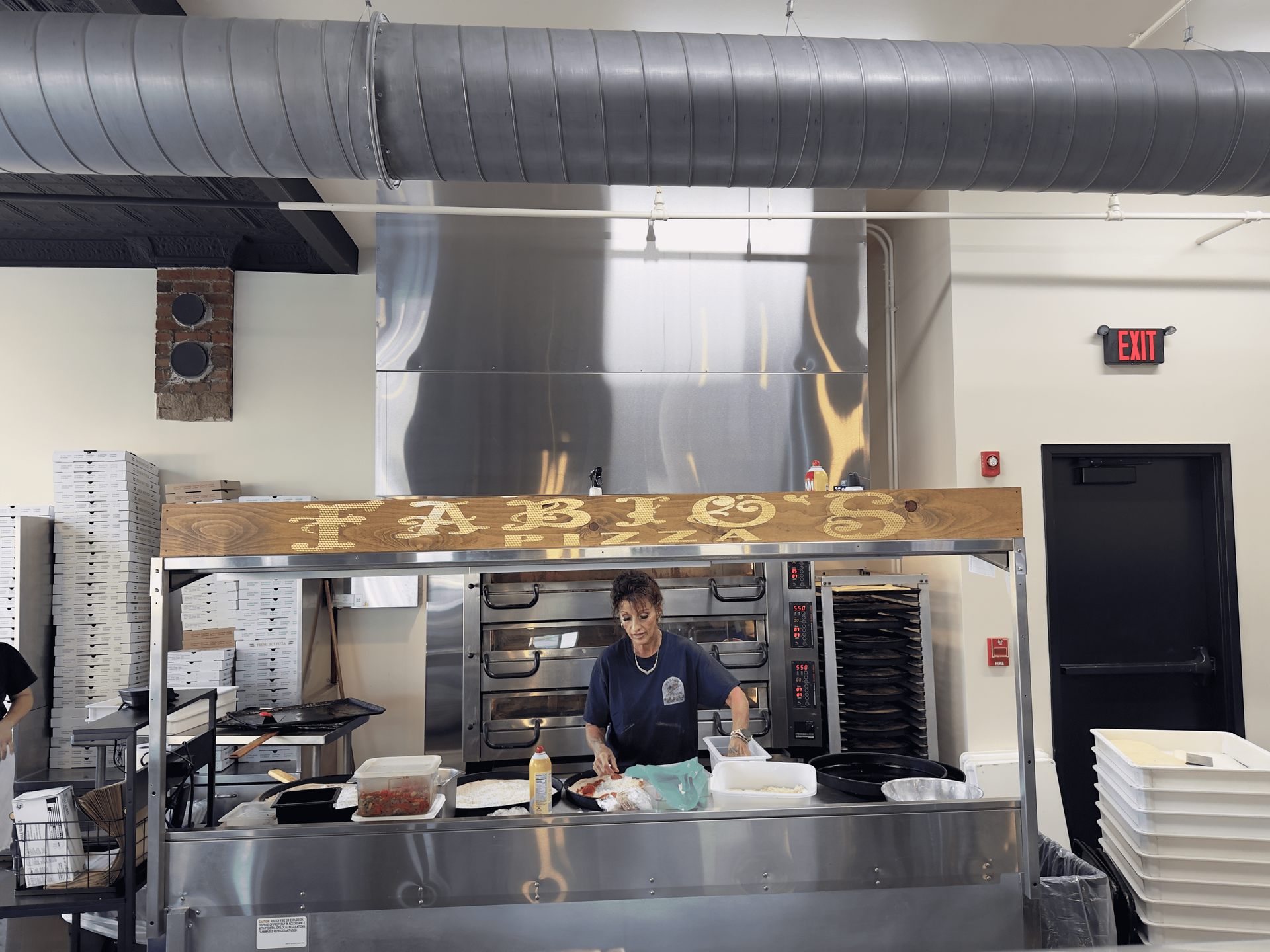 Restaurant kitchen with a cook preparing food at a stainless steel counter under a large ventilation hood.