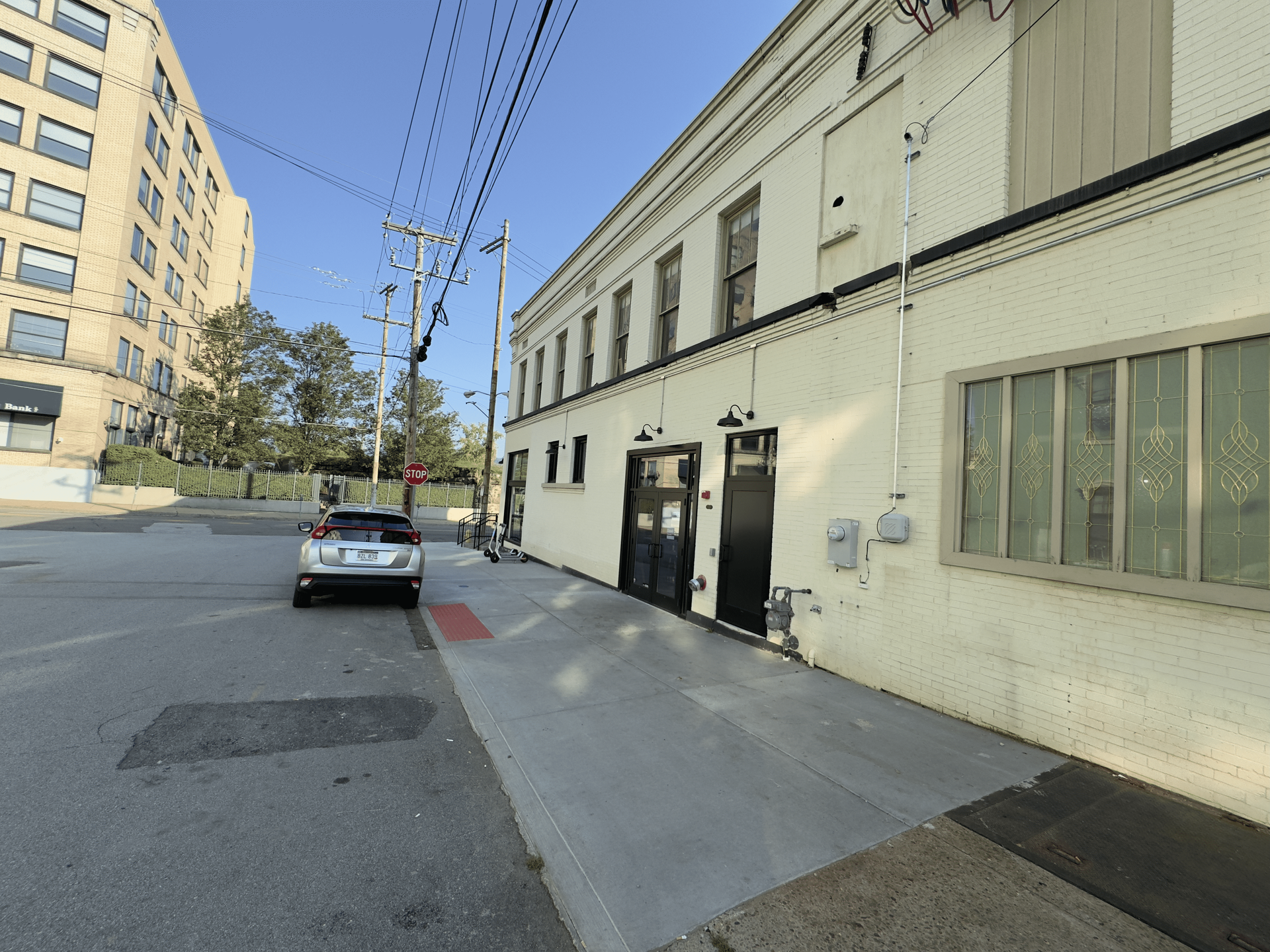Quiet urban street beside a beige building, with a parked car, sidewalk, and overhead power lines under a clear blue sky