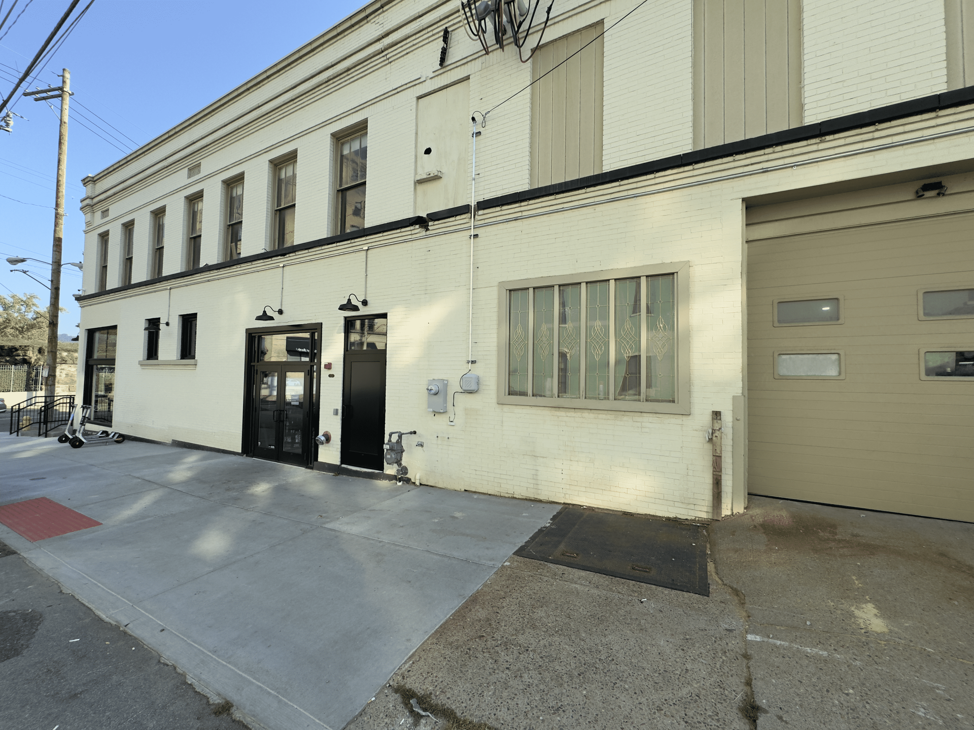 Exterior of a light-colored building with a black door, garage door, and sidewalk on a sunny street