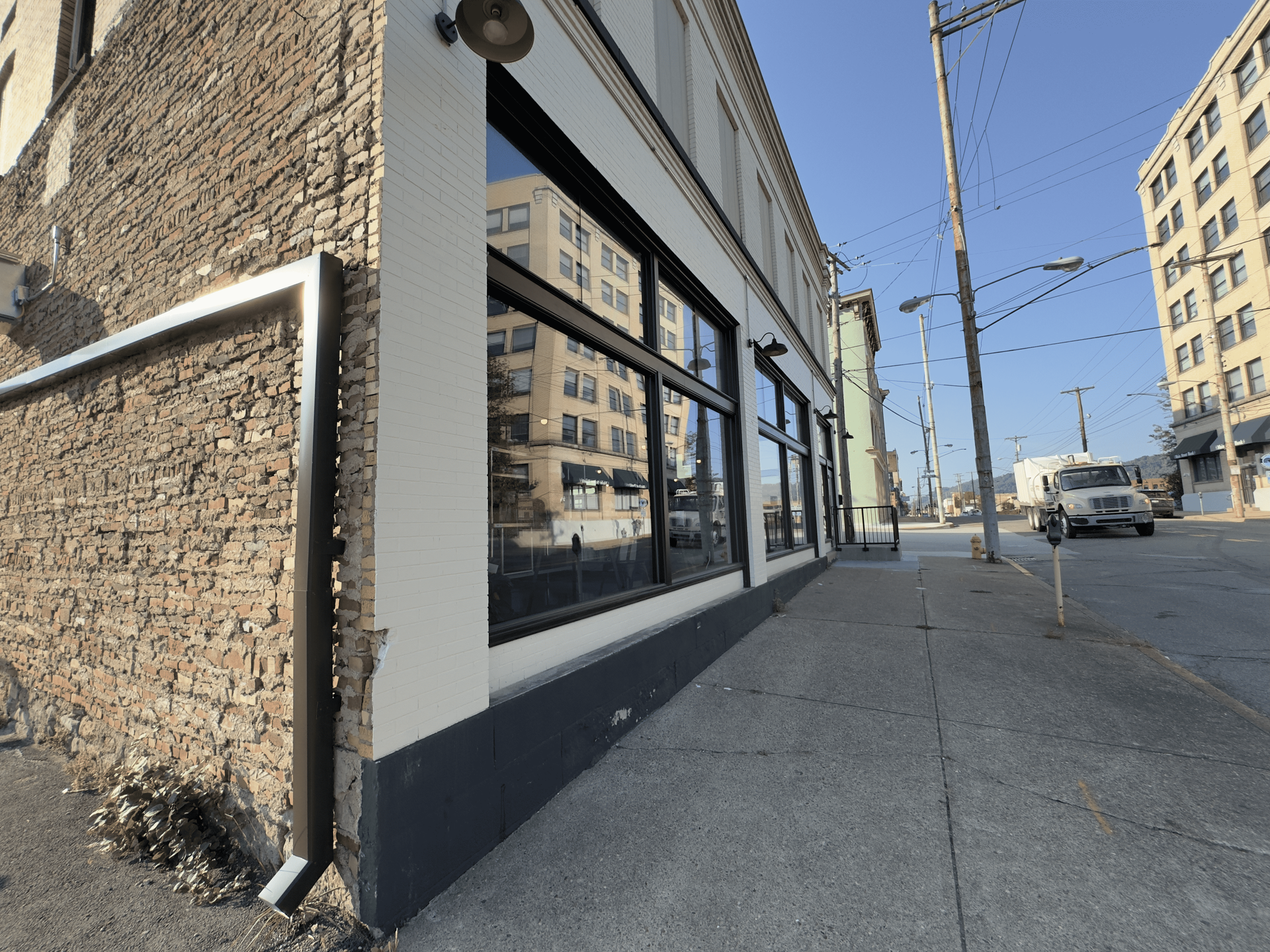 Urban sidewalk beside a brick building with large windows, parked cars, and a clear blue sky