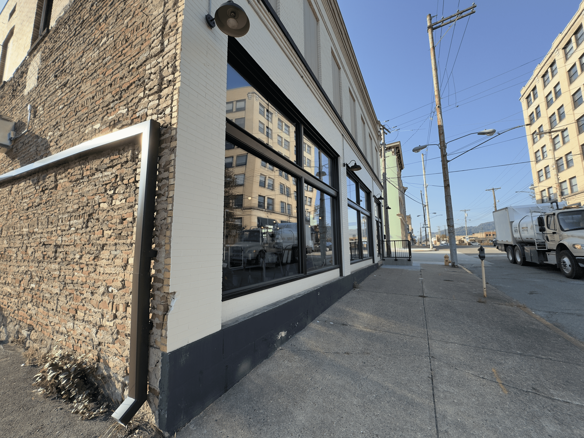 Corner storefront on a sunny street, with large windows, brick walls, and parked trucks nearby.