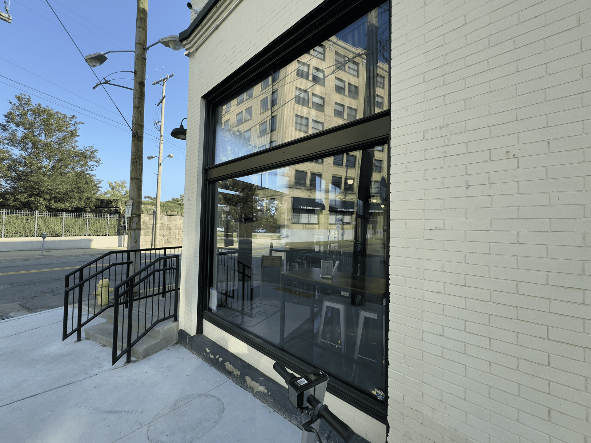 Exterior of a storefront with large black-framed windows, a ramp, and a white brick wall on a sunny street.