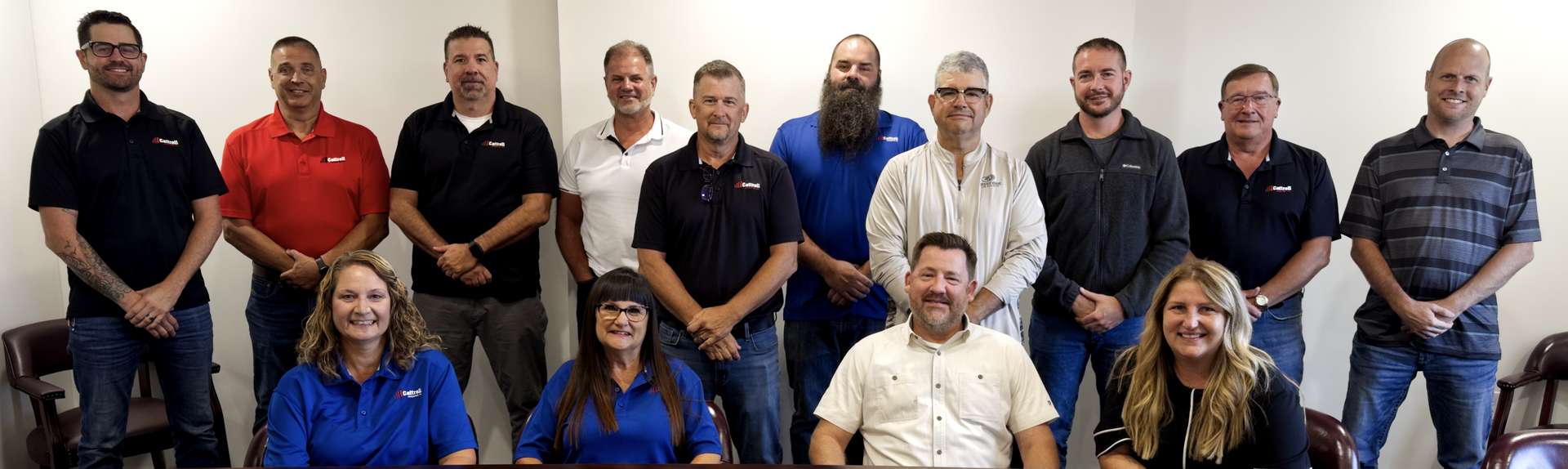 Group of coworkers posing indoors, standing and sitting in two rows against a plain wall.