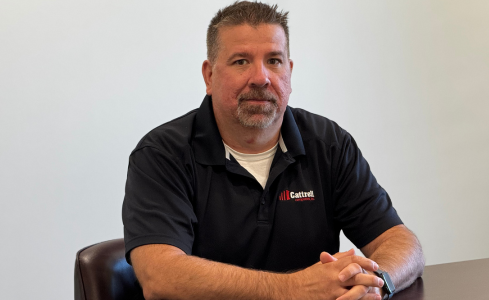 Man in a black polo shirt seated at a table, facing the camera in an office setting.