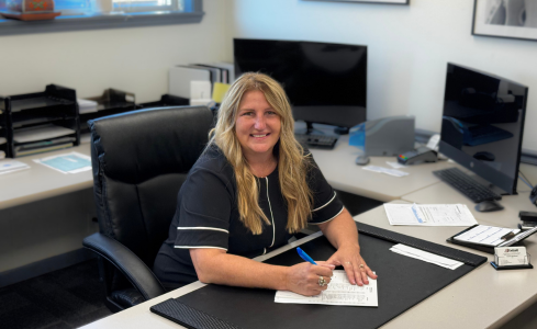 Smiling person at office desk writing on paper, with computers and paperwork nearby.