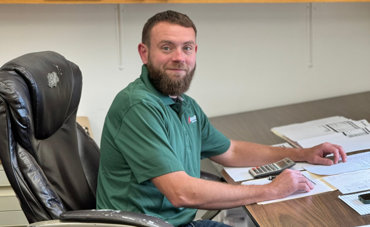 Smiling man in black polo shirt seated at a table in an office setting