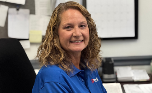 Smiling woman in a blue polo shirt at a desk in an office.